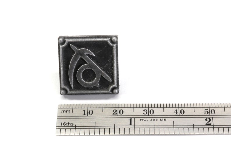 May include: A silver metal pin with a stylized symbol of a sickle and a circle. The pin is about 1.5 inches wide and is shown next to a ruler for scale.