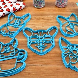 Pu&ograve; includere: Set di cinque tagliabiscotti in plastica blu a forma di corgi. I tagliabiscotti presentano diverse espressioni di corgi, tra cui uno con gli occhiali e uno che tiene una tazza.
