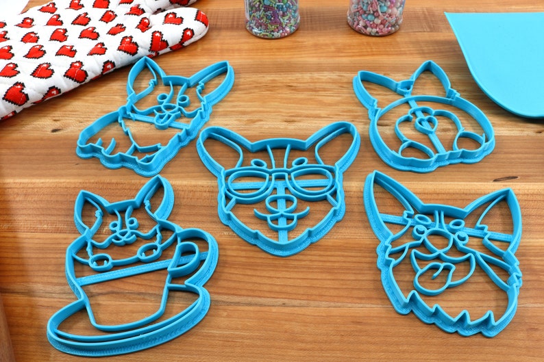 Pu&ograve; includere: Set di cinque tagliabiscotti in plastica blu a forma di corgi. I tagliabiscotti presentano diverse espressioni di corgi, tra cui uno con gli occhiali e uno che tiene una tazza.
