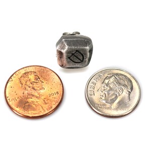 May include: A silver metal dice with a letter D engraved on one side. The dice is sitting on a white surface next to a US penny and a US dime.