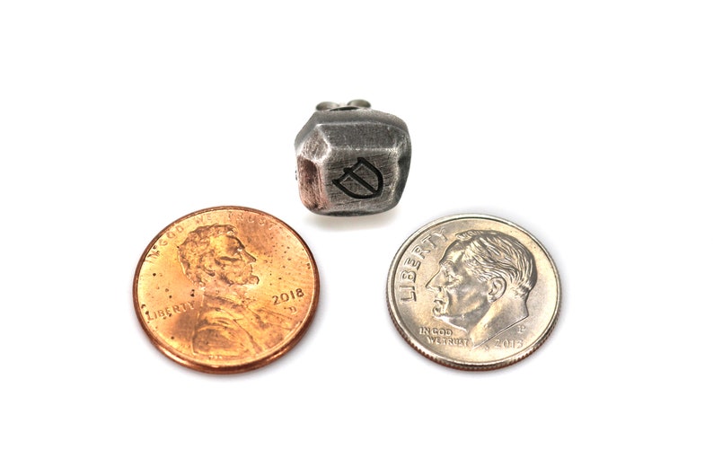 May include: A silver metal dice with a letter D engraved on one side. The dice is sitting on a white surface next to a US penny and a US dime.