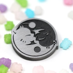 May include: A silver and black yin yang symbol with a stylized cat and bat design. The symbol is a circular pendant with a raised edge.