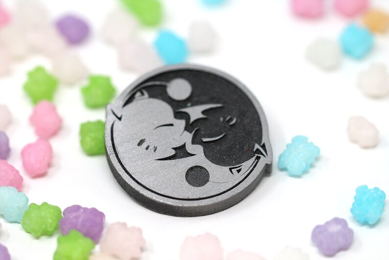 May include: A silver and black yin yang symbol with a stylized cat and bat design. The symbol is a circular pendant with a raised edge.