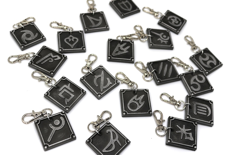 May include: A set of 16 silver keychains with different symbols on each. The keychains are black with silver accents and are all square shaped.
