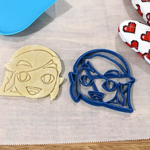 May include: Two cookie cutters, one blue and one beige, shaped like a cartoon character with a headband and large eyes. The cutters are on a white parchment paper surface.
