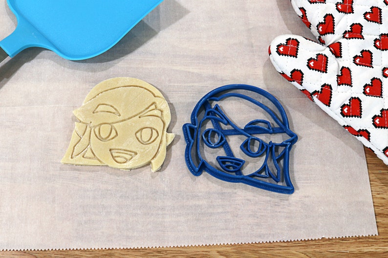 May include: Two cookie cutters, one blue and one beige, shaped like a cartoon character with a headband and large eyes. The cutters are on a white parchment paper surface.