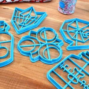 May include: Six blue plastic cookie cutters on a wooden surface. The cutters are shaped like a bottle, a geometric shape, a circle with a design inside, and a jar.
