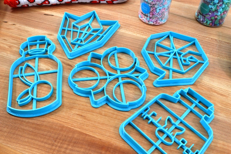 May include: Six blue plastic cookie cutters on a wooden surface. The cutters are shaped like a bottle, a geometric shape, a circle with a design inside, and a jar.