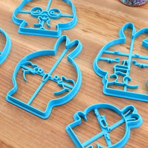 May include: Five blue plastic cookie cutters shaped like cartoon characters. The cutters are on a wooden surface.