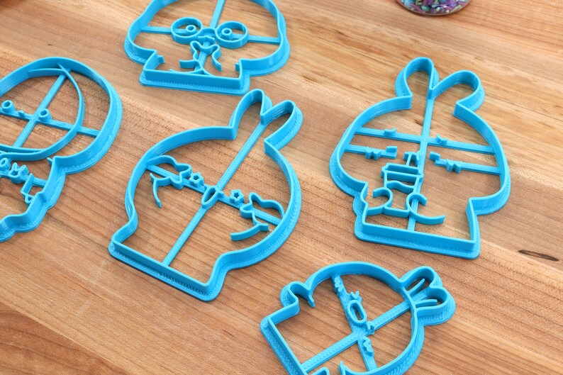 May include: Five blue plastic cookie cutters shaped like cartoon characters. The cutters are on a wooden surface.