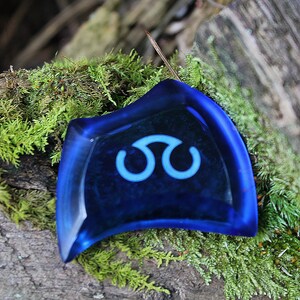 May include: A blue glass pendant with a white symbol resembling a stylized infinity sign. The pendant is resting on a bed of green moss.