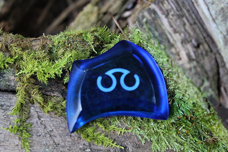 May include: A blue glass pendant with a white symbol resembling a stylized infinity sign. The pendant is resting on a bed of green moss.