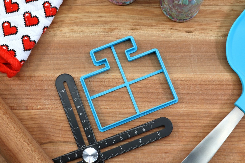 May include: A blue plastic cookie cutter shaped like a house with four windows. The cutter is on a wooden surface with a ruler and other baking tools.