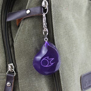 Op de afbeelding: Een paarse sleutelhanger in de vorm van een traan met een gestileerd vlamontwerp erin. De sleutelhanger is bevestigd aan een zilveren metalen ring en hangt aan een groene canvas tas.