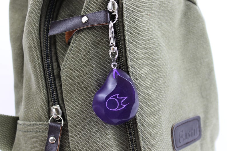 Op de afbeelding: Een paarse sleutelhanger in de vorm van een traan met een gestileerd vlamontwerp erin. De sleutelhanger is bevestigd aan een zilveren metalen ring en hangt aan een groene canvas tas.