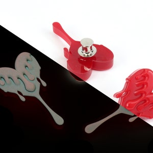 May include: Two red heart-shaped pins with dripping edges. One pin has a silver back with a pin post. The other pin is flat.