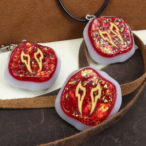 May include: Three red resin pendants with gold accents in the shape of flames. The pendants are on a brown leather surface. One pendant is attached to a black cord, another is attached to a silver key chain, and the third pendant is loose.