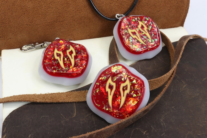 May include: Three red resin pendants with gold accents in the shape of flames. The pendants are on a brown leather surface. One pendant is attached to a black cord, another is attached to a silver key chain, and the third pendant is loose.