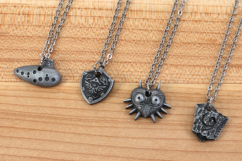 May include: Four silver necklaces with different pendants: an ocarina, a shield with a triforce symbol, a Majora's Mask, and a stone with a swirl design.