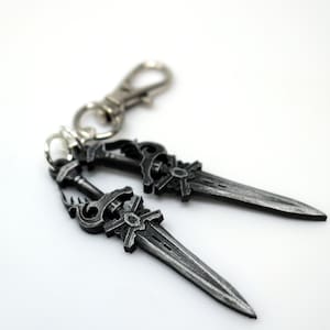 May include: A silver-colored keychain featuring two miniature dagger pendants. The daggers have detailed handles and blades, connected to a clasp. The design has an antique or fantasy aesthetic.