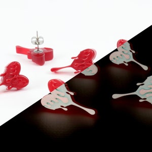 May include: A pair of red heart-shaped earrings with a dripping effect. The earrings are shown in both natural and blacklight conditions, revealing a pink and green glow under blacklight.