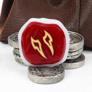 May include: A red and white gaming die with a yellow symbol of two flames. The die is sitting on top of a stack of silver coins.