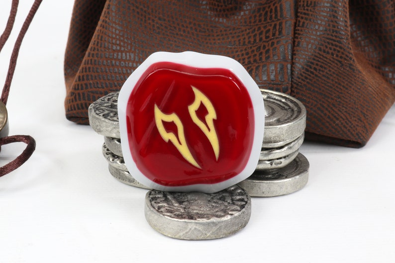 May include: A red and white gaming die with a yellow symbol of two flames. The die is sitting on top of a stack of silver coins.