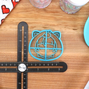 Pu&ograve; includere: Tagliapasta in plastica blu a forma di personaggio dei cartoni animati con orecchie da gatto e papillon. Il tagliapasta &egrave; su una superficie di legno con un righello e una spatola blu.