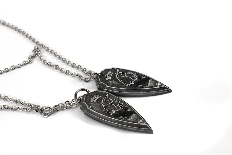 May include: Two silver necklaces with matching pendants. Each pendant is shaped like a shield with a detailed design of a mythical creature.