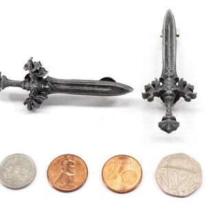 May include: Two silver-colored sword pins with intricate designs. The pins are shown next to four coins for size comparison.