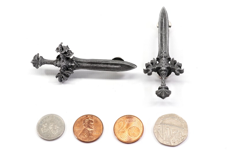 May include: Two silver-colored sword pins with intricate designs. The pins are shown next to four coins for size comparison.