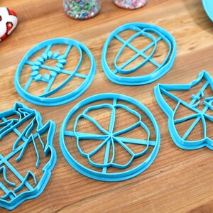 May include: Five blue plastic cookie cutters on a wooden surface. The cutters are shaped like a flower, a sun, an egg, a leaf, and a shoe.