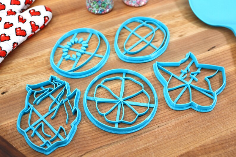 May include: Five blue plastic cookie cutters on a wooden surface. The cutters are shaped like a flower, a sun, an egg, a leaf, and a shoe.
