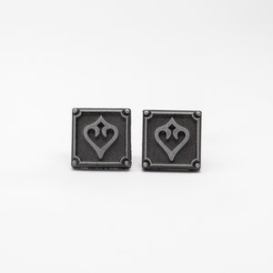 May include: A pair of silver-toned metal cufflinks with a square design featuring a stylized heart in the center.