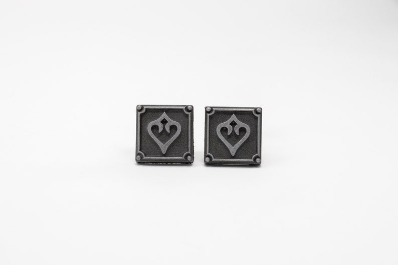 May include: A pair of silver-toned metal cufflinks with a square design featuring a stylized heart in the center.