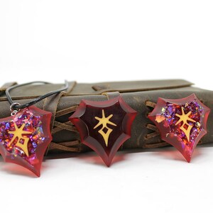May include: Three red star-shaped pendants with gold symbols inside. The pendants are on a brown leather journal with a black cord.