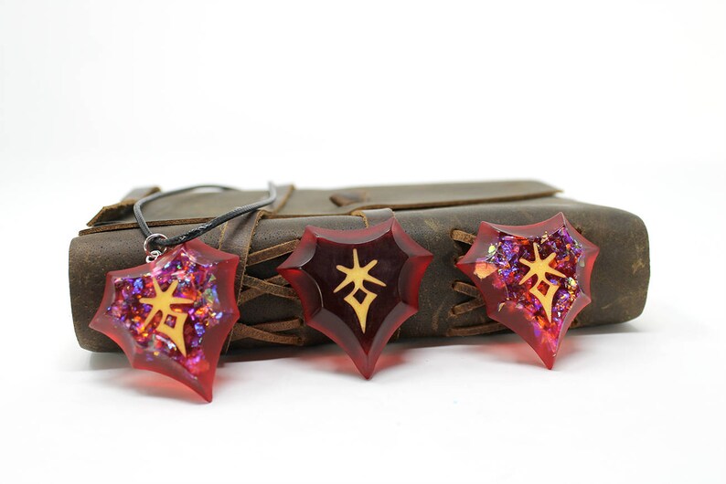 May include: Three red star-shaped pendants with gold symbols inside. The pendants are on a brown leather journal with a black cord.