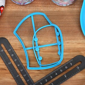 May include: A blue plastic cookie cutter shaped like a chef's hat with a grid pattern and five small circles. The cutter is on a wooden surface with two black rulers.