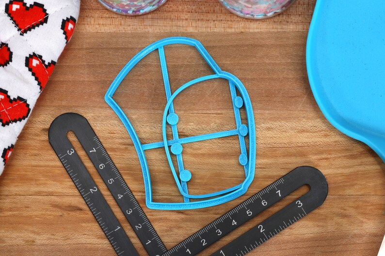 May include: A blue plastic cookie cutter shaped like a chef's hat with a grid pattern and five small circles. The cutter is on a wooden surface with two black rulers.