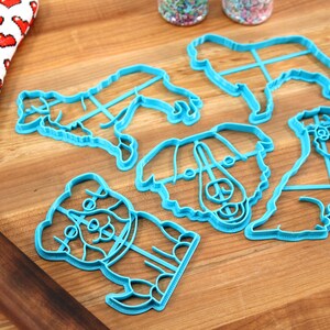 May include: A set of six blue plastic dog-shaped cookie cutters on a wooden surface. The cutters are different sizes and shapes, and some have detailed features like eyes and noses.