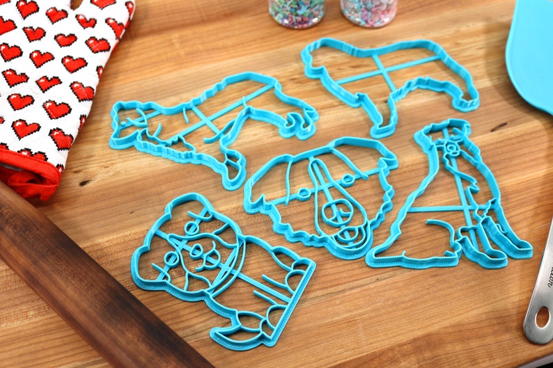May include: A set of six blue plastic dog-shaped cookie cutters on a wooden surface. The cutters are different sizes and shapes, and some have detailed features like eyes and noses.