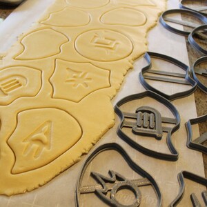 Op de afbeelding: Een vel koekdeeg met verschillende vormen uitgesneden, waaronder een cirkel met een symbool erin, een schild met een symbool erin en een halve maan met een symbool erin. Naast het deeg is een set koekjesvormen met dezelfde vormen en symbolen.