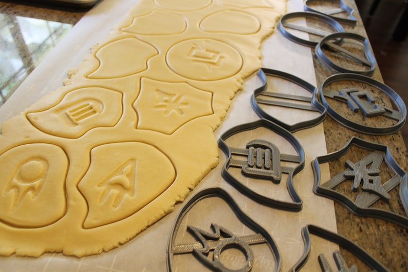 Op de afbeelding: Een vel koekdeeg met verschillende vormen uitgesneden, waaronder een cirkel met een symbool erin, een schild met een symbool erin en een halve maan met een symbool erin. Naast het deeg is een set koekjesvormen met dezelfde vormen en symbolen.