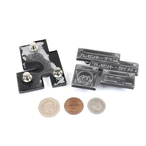 Op de afbeelding: Twee zilverkleurige metalen pinnen met een zwart-wit ontwerp. De pinnen zijn gevormd als computervensters met tekst die "Alert -  Sterf" en "Je lijkt niet te begrijpen... Een aandeel" luidt.  De linker pin heeft vier zilverkleurige metalen knoppen.  Drie Nederlandse munten bevinden zich in het midden onderaan de afbeelding.