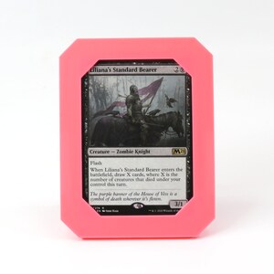 Op de afbeelding: Een zwart-witte Magic: The Gathering kaart met een roze rand. De kaart toont een zombie-ridder die een paard met een paarse vlag berijdt. De tekst op de kaart luidt "Liliana's Standard Bearer" en "Creature - Zombie Knight".