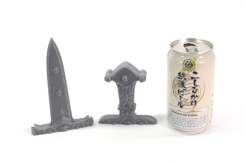 May include: Three gray 3D printed miniature swords, one with a long blade and two with shorter blades. The swords are on a white surface. A can of Japanese beer is in the background.