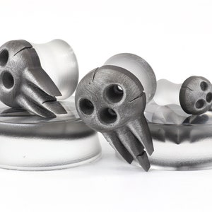 May include: A set of gray, decorative ear plugs with a unique design. Each plug features a stylized, three-holed face with pointed features. The plugs are set on clear, cylindrical bases, creating a modern, artistic aesthetic.