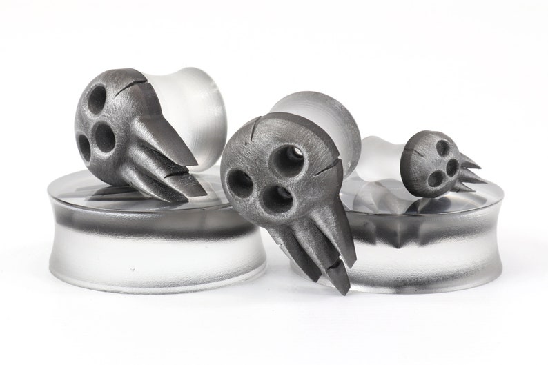 May include: A set of gray, decorative ear plugs with a unique design. Each plug features a stylized, three-holed face with pointed features. The plugs are set on clear, cylindrical bases, creating a modern, artistic aesthetic.