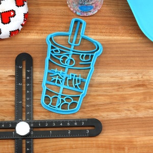 May include: A light blue plastic cookie cutter in the shape of a bubble tea cup with Chinese characters. The cutter is on a wooden surface with other items, including a black measuring tool and a heart-patterned item.