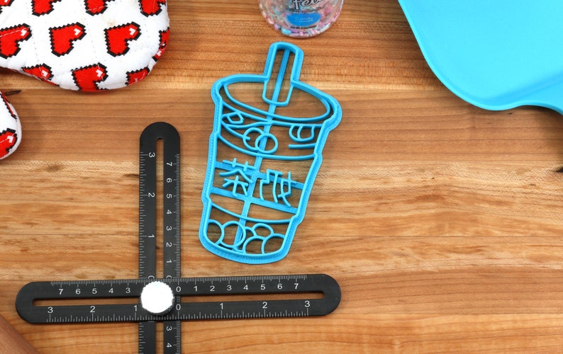May include: A light blue plastic cookie cutter in the shape of a bubble tea cup with Chinese characters. The cutter is on a wooden surface with other items, including a black measuring tool and a heart-patterned item.